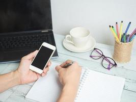 mão segurando o smartphone e escrevendo no espaço de trabalho do notebook no fundo da mesa de madeira branca foto