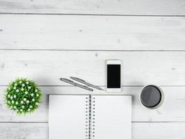 espaço de trabalho de escritório espaço de cópia de visão superior, tela preta do smartphone no caderno de página preta com óculos e duas canetas de prata na mesa de madeira branca foto