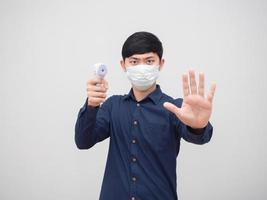 homem asiático usando máscara mostra parada de mão e segurando termômetro infravermelho na mão cara séria no fundo branco foto