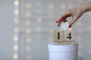 conceito de dia dos namorados. data 14 de fevereiro no calendário do cubo de madeira em uma caixa de presente branca e coração branco na mão. bokeh fundo dourado e branco. foto