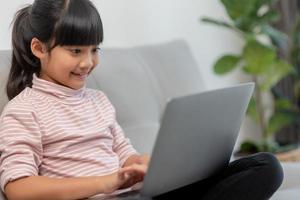 garotinha asiática da escola usa computador portátil sentado no sofá sozinho em casa. criança aprendendo lendo conteúdo de mídia social on-line, jogando lições de educação conversando com amigos. foto