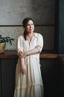 retrato de uma mulher sorridente morena de meia-idade adulta amigável cinquenta anos em vestido branco na cozinha com plantas de casa verde em casa foto