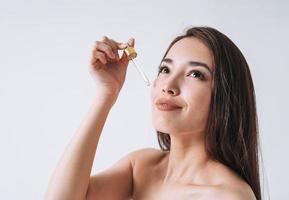 retrato da beleza da mulher asiática sorridente feliz com cabelos longos escuros com pipeta com óleo orgânico no rosto de pele fresca limpa e mãos sobre fundo branco isolado foto