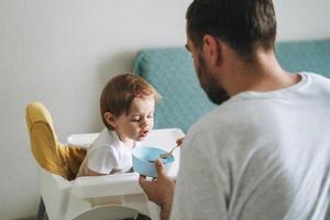 feliz pai jovem alimenta bebê filhinha na cozinha em casa foto