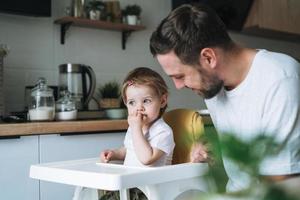 feliz pai jovem alimenta bebê filhinha na cozinha em casa foto