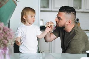 jovem pai feliz alimentando e se divertindo com a filhinha na cozinha em casa foto