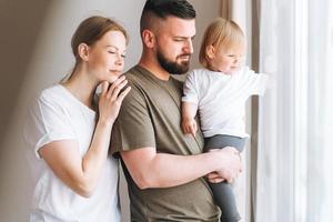 feliz família multinacional pai jovem e mãe com filha pequena se divertindo perto da sala da janela em casa foto