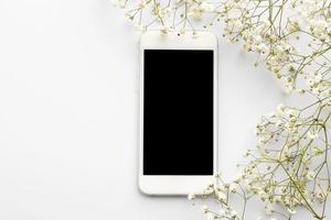 celular branco e flores em um fundo branco. um smartphone com uma tela em branco. A vista do topo. conceito de verão primavera. copie o espaço para design foto