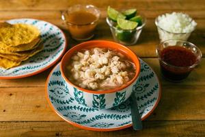 pozole servido em prato fundo, com tostadas, salsa e legumes em mesa de madeira. comida típica mexicana. foto