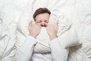 homem de pijama branco está estressado, tem insônia, não consegue dormir, cobre nervosamente a boca com um travesseiro e fecha os olhos com força. vista do topo. foto