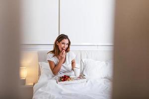 jovem sorridente tomando café da manhã saboroso e café em uma bandeja na cama. mulher tomando café da manhã na cama. grande sorriso cheio de dentes. foto