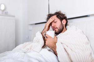 dia doente em casa. homem bonito tem febre e resfriado comum. tosse. closeup de jovem pegou resfriado ou gripe. retrato de homem insalubre sentindo dor na garganta e bebendo bebida quente. foto