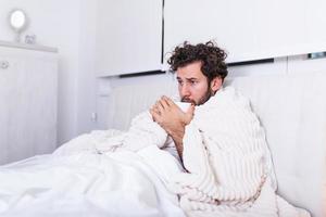 dia doente em casa. homem bonito tem febre e resfriado comum. tosse. closeup de jovem pegou resfriado ou gripe. retrato de homem insalubre sentindo dor na garganta e bebendo bebida quente. foto