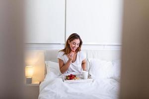 mulher atraente feliz acordou de manhã na cama. mulher tomando café da manhã no quarto de hotel, mulher tomando café da manhã na cama com frutas, café e biscoitos foto