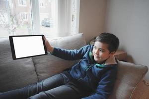 criança feliz com computador tablet. garoto de retrato segurando e mostrando tablet digital com maquete de tela vazia, jovem sentado no sofá relaxando e brincando com o touch pad em casa foto