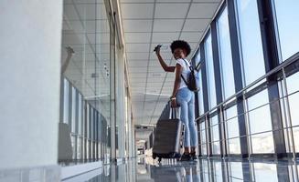 vista traseira se jovem passageira afro-americana em roupas casuais que está no aeroporto com bagagem foto