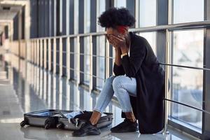 triste e deprimida jovem passageira afro-americana em roupas casuais, sentada no aeroporto com bagagem foto