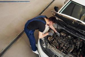 sob o capô. homem de uniforme de trabalho repara automóvel branco dentro de casa. concepção de serviço automóvel foto