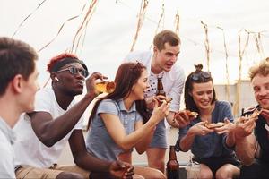 com deliciosas pizzas. grupo de jovens em roupas casuais faz uma festa no telhado juntos durante o dia foto