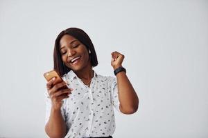 mulher afro-americana positiva na camisa branca em pé dentro de casa contra a parede branca com o telefone na mão foto