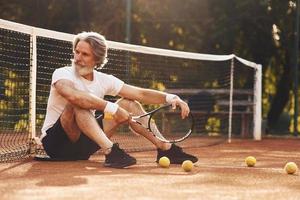 sentado e fazendo uma pausa. homem estiloso sênior em camisa branca e calção esportivo preto na quadra de tênis foto