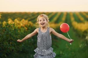 menina positiva com balão vermelho nas mãos divirta-se no campo durante o dia de verão foto