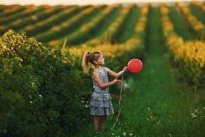 menina positiva com balão vermelho nas mãos divirta-se no campo durante o dia de verão foto