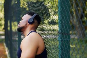 cara afro-americano em fones de ouvido sem fio se inclina sobre a malha de metal da quadra esportiva e faz uma pausa foto