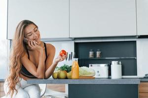 fala ao telefone. jovem europeia está dentro de casa na cozinha dentro de casa com comida saudável foto