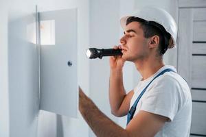 jovem eletricista masculino no capacete branco trabalha dentro de casa na sala foto