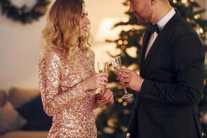 de pé contra a árvore de natal. casal feliz tem uma festa de ano novo dentro de casa juntos foto