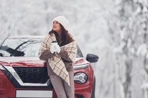 na frente do carro dela. bela jovem está ao ar livre perto de seu automóvel vermelho no inverno foto