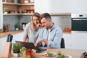 olhando para portátil. casal preparando comida em casa na cozinha moderna foto