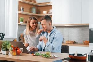 olhando para portátil. casal preparando comida em casa na cozinha moderna foto