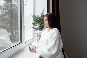 mulher de suéter de inverno branco quente ao lado da janela em casa na véspera de Natal segurando uma xícara com marshmallows, abeto atrás foto