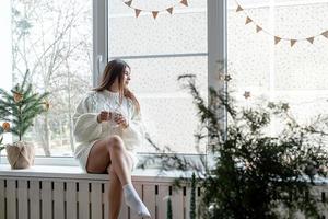 mulher com suéter de inverno branco quente sentado no parapeito da janela em casa na véspera de natal segurando copo com marshmallows foto