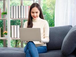 mulheres asiáticas bonitas e atraentes ficam em casa com um laptop fazendo trabalhos de pesquisa para seus negócios enquanto estão sentadas no sofá da sala de estar. foto