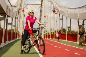 linda garota com cabelo loiro andando de bicicleta alegremente. retrato de jovem em j óculos de sol se divertindo, andando de bicicleta pelas ruas da cidade foto