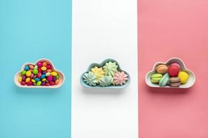 doces coloridos - pirulitos, merengues, biscoito em tigela em forma de nuvem isolado em rosa, branco, fundo azul plano de visão superior knolling comida insalubre e saborosa conceito criativo cartão de férias foto