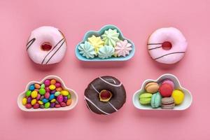 doces e merengues em tigela em forma de nuvem, chocolate com cobertura colorida e rosquinha rosa isolada em fundo rosa plana vista de cima knolling conceito de comida insalubre e saborosa foto