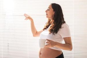 curiosa grávida bebendo chá e olhando pela janela pelas persianas. conceito de alegria e boas notícias enquanto espera pelo bebê foto