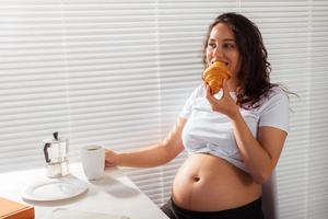 mulher bonita jovem grávida feliz comendo croissant durante o café da manhã. conceito de manhã agradável e atitude positiva durante a gravidez foto