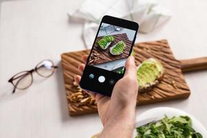 as mãos tiram fotos no smartphone de dois lindos sanduíches de creme azedo e abacate a bordo sobre a mesa. mídia social e conceito de comida