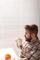 Vista lateral do pensativo jovem empresário masculino barbudo bebendo café no fundo das persianas. conceito de manhã agradável ou pausa para o almoço. foto