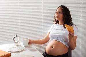 mulher bonita jovem grávida feliz comendo croissant durante o café da manhã. conceito de manhã agradável e atitude positiva durante a gravidez foto