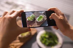 as mãos tiram fotos no smartphone de dois lindos sanduíches de creme azedo e abacate a bordo sobre a mesa. mídia social e conceito de comida