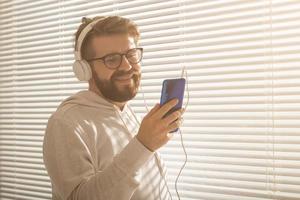 retrato de close-up do homem jovem elegante hipster escolhendo música favorita em smartphone com fones de ouvido no escritório. conceito de assinatura de música foto