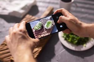 as mãos tiram fotos no smartphone de dois lindos sanduíches de creme azedo e abacate a bordo sobre a mesa. mídia social e conceito de comida