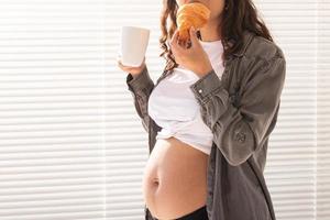 mulher grávida bonita saudável bebendo chá e comendo croissant durante o almoço. conceito de nutrição de alto teor calórico enquanto aguarda o nascimento do bebê. copie o espaço. foto