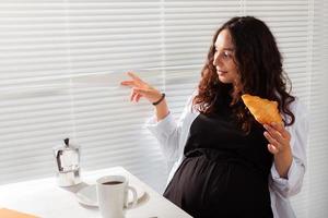 mulher bonita jovem grávida feliz comendo croissant e olhando através das persianas durante o café da manhã. conceito de manhã agradável e atitude positiva durante a gravidez foto